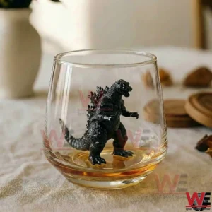 Whiskey glass with a black Godzilla figure inside surrounded by a small amount of amber whiskey on a neutral tabletop background!