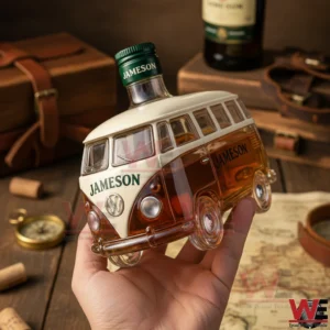 A hand holding a camper van shaped whiskey bottle filled with brown liquid displayed on a wooden table with maps leather cases and vintage travel items!