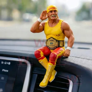 Miniature wrestler figure in yellow and red outfit sitting on a car dashboard with a championship belt and a hand raised to the ear.