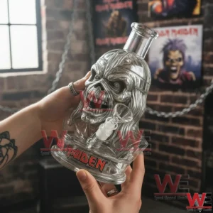Iron Maiden Eddie Bong 4 Hands holding a clear glass skull water pipe with Iron Maiden logo against a brick wall with Iron Maiden posters.