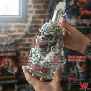Iron Maiden Eddie Bong 5 Clear glass skull shaped water pipe with Iron Maiden logo held in both hands in front of brick wall and band posters.