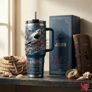 Blue handled tumbler with embossed shark and ocean themed details beside a matching Jaws gift box on a rustic table with rope anchor shells compass and books!