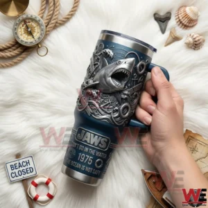 Hand holding a blue handled tumbler with embossed shark and ocean wave details on white fur surrounded by rope compass shells map beach closed sign and nautical props!