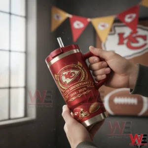 Hands holding a red Kansas City Chiefs tumbler with red lid and clear straw featuring the arrowhead logo and Sixty Seventh Anniversary Nineteen Fifty Nine to Two Thousand Twenty Six design in a team decorated room.