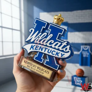 A hand holding a Kentucky Wildcats whisky bottle with the UK logo blue glass accents amber liquid and a gold crown stopper in a sports themed setting!