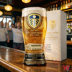 A golden beer filled Leeds United anniversary glass on a bar counter with fans celebrating in the background and a matching box placed nearby!