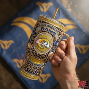 A hand holds a Los Angeles Rams insulated tumbler decorated with ornate design elements team logo and historical details against a soft indoor backdrop!