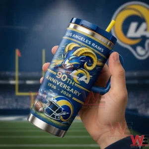 Hand holding a blue Los Angeles Rams tumbler with navy lid and yellow straw featuring a ram head graphic and Ninety Th Anniversary Nineteen Thirty Six to Two Thousand Twenty Six design in front of a stadium background.