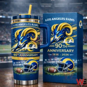 Blue Los Angeles Rams tumbler with navy lid and yellow straw on a wooden table next to a matching collector box featuring a ram head graphic and Ninety Th Anniversary Nineteen Thirty Six to Two Thousand Twenty Six design in front of a stadium crowd.