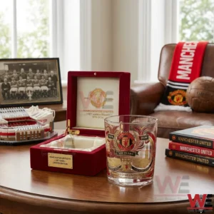 A Manchester United anniversary commemorative glass sitting on a wooden table with a red presentation box books and club themed decorations in a stylish room!