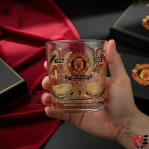 A hand holding a Manchester United commemorative glass with intricate anniversary design set against a red fabric background!