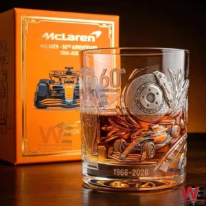 A McLaren 60th anniversary whiskey glass with racing car engraving brake disc detail and amber whiskey displayed in front of an orange commemorative box!