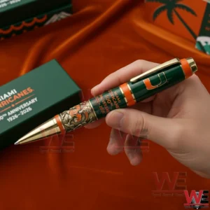 Hand holding a Miami Hurricanes 100th anniversary 1926 2026 commemorative pen in green and orange with gold trim and mascot artwork with a Miami Hurricanes gift box in the background!
