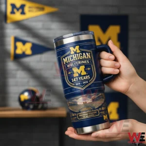 Hands holding a navy Michigan Wolverines tumbler with a handle and clear lid featuring the block M logo One Hundred Forty Seven Years Eighteen Seventy Nine to Two Thousand Twenty Six and a Michigan Stadium The Big House illustration in a room with team banners.