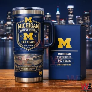 Navy Michigan Wolverines tumbler with handle and clear lid on a wooden table next to a matching limited edition box featuring the block M logo and One Hundred Forty Seven Years Eighteen Seventy Nine to Two Thousand Twenty Six design with a city skyline background.
