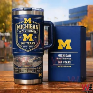 Navy Michigan Wolverines tumbler with handle and clear lid on a wooden table next to a matching limited edition box featuring the block M logo One Hundred Forty Seven Years Eighteen Seventy Nine to Two Thousand Twenty Six and Michigan Stadium The Big House artwork in an outdoor campus setting.