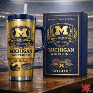 Gold Michigan Wolverines collector edition insulated tumbler with handle and straw beside a matching anniversary box on a wooden table with a blurred stadium background!
