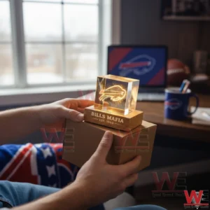 Hands holding a warm glowing crystal cube lamp with an etched football logo on a wooden base engraved Bills Mafia in a cozy sports themed room.