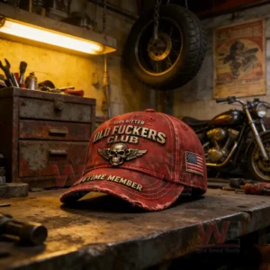 Distressed red baseball cap with embroidered skull wings emblem and bold text on a worn workbench in a rustic motorcycle garage with tools and a hanging tire in the background!