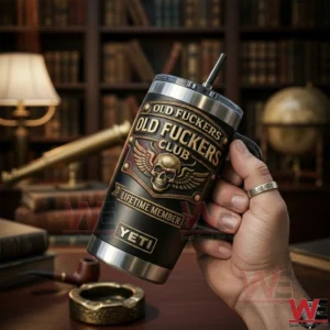 Hand holding a black and gold tumbler with Old Fuckers Club Lifetime Member winged skull emblem and metal straw in a vintage library background!