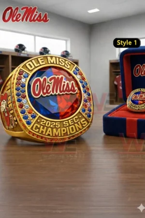 Gold Ole Miss 2025 SEC Champions ring with red and blue gemstone style face Ole Miss logo red blue and clear stones and a matching display box on a wooden table in a locker room setting!