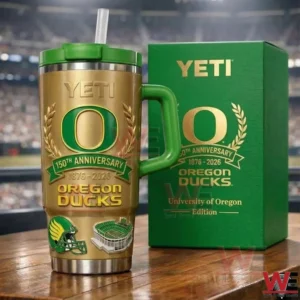 Gold Oregon Ducks insulated tumbler with green handle lid and straw featuring anniversary branding and team graphics displayed on a table with a stadium background!