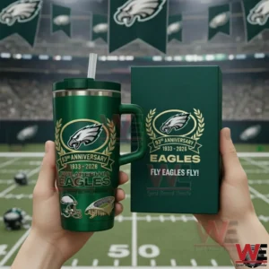 Hands holding a green Philadelphia Eagles tumbler with a straw next to a matching collector box featuring the Eagles logo Ninety Third Anniversary Nineteen Thirty Three to Two Thousand Twenty Six and Fly Eagles Fly text in a stadium setting.