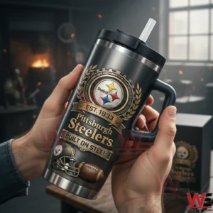 Hands holding a dark Pittsburgh Steelers tumbler with handle black lid and clear straw featuring the Steelers logo Est Nineteen Thirty Three Pittsburgh Steelers Built On Steel and football icons in a warm indoor room.