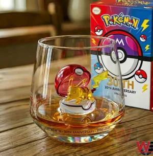A Pokemon anniversary whiskey glass with a Pikachu figure and themed packaging displayed on a wooden surface!