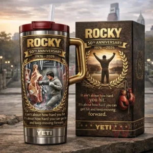 Dark brown insulated tumbler with gold handle and straw featuring Rocky fiftieth anniversary boxing artwork beside a matching collector box on a stone surface with a city skyline background!