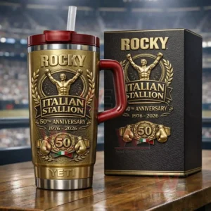 Gold Rocky Italian Stallion 50th anniversary tumbler with red handle lid and straw embossed boxer emblem 1976 2026 text and matching collector box on a wooden table with stadium background