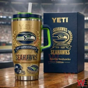 Gold Seattle Seahawks fiftieth anniversary insulated tumbler with navy and green handle and straw lid next to a matching navy limited edition gift box on a wooden table with a blurred stadium background!
