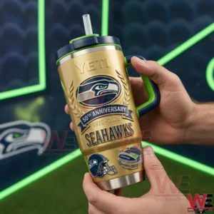 Hands holding a gold Seattle Seahawks tumbler with navy lid clear straw and green handle featuring the Seahawks logo Fiftieth Anniversary Nineteen Seventy Six to Two Thousand Twenty Six and football icons in a stadium themed background.