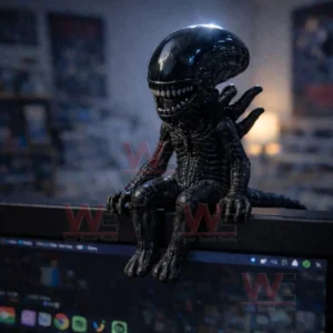 Miniature black alien creature figurine with elongated head and sharp teeth sitting on the top edge of a computer monitor in a dim room