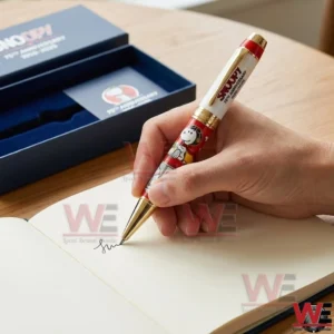 Snoopy 75th Anniversary 1950-2025 Pen 5 Hand writing in a notebook with a Snoopy 75th anniversary pen while an open navy gift box and anniversary card appear in the background!