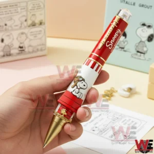 Hand holding a Snoopy commemorative pen with red white body gold accents and Snoopy artwork in a cute themed background.