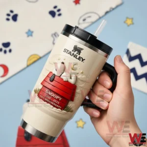 Snoopy Sleep on a Roof 75Th Anniversary Stanley Tumbler Cup 5 Hand holding a cream Stanley tumbler with black handle and straw featuring Snoopy 75th anniversary artwork on a red doghouse!