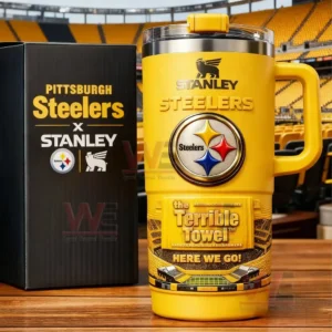 Yellow Stanley Steelers tumbler with handle Steelers logo Terrible Towel Here We Go text and matching Pittsburgh Steelers x Stanley box on a wooden table with stadium seating background