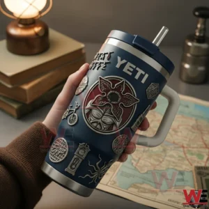 Hand holding a navy Yeti tumbler with a handle and textured emblem details on a desk with books and a map!
