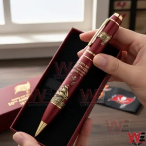 Hand holding a Tampa Bay Buccaneers 50th anniversary 1976 2026 commemorative pen in a maroon presentation box featuring pirate flag branding and a ship illustration with team themed items behind!