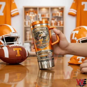 Hand holding an orange Tennessee Volunteers 135 years insulated tumbler with handle and straw next to a football and team helmets on a sports themed table.