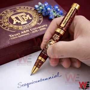 Hand writing with a Texas AM 150th anniversary maroon and gold pen while a matching gift box and blue flowers appear behind!