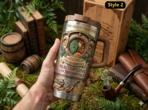 The Hobbit The Shire Bag End 2001-2026 25th Anniversary Tumbler 3 Hand holding a Bag End Shire themed insulated tumbler with twenty fifth anniversary text and wood style accents surrounded by books and greenery.