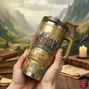 Hands holding a gold fantasy themed tumbler with handle and straw featuring a fellowship illustration One Journey One Oath Two Thousand One to Two Thousand Twenty Six and Twenty Fifth Anniversary design on a table with books and a mountain valley backdrop.