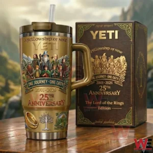 Gold fantasy themed tumbler with handle and straw on a wooden table beside a matching collector box featuring fellowship artwork One Journey One Oath Two Thousand One to Two Thousand Twenty Six and Twenty Fifth Anniversary design with a mountain valley background.