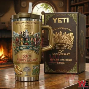 Gold fantasy themed tumbler with handle and straw on a wooden table next to a matching collector box featuring fellowship artwork One Journey One Oath Two Thousand One to Two Thousand Twenty Six and Twenty Fifth Anniversary design in a cozy room with a fireplace.