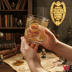 The Lord of the Rings Fellowship of the Nine 25th Anniversary Whiskey Glass 3 Hands holding a The Lord Of The Rings whiskey glass with gold elvish artwork ring design and 25th anniversary 2001 2026 text in a cozy fantasy themed room with books and figurines
