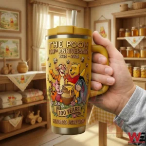 The Pooh 100th Anniversary 100 Years Hundred Acre Wood Tumbler 5 Hand holding a yellow The Pooh 100th Anniversary insulated tumbler with handle and straw featuring classic characters honey pot artwork and anniversary dates with a cozy indoor themed background!