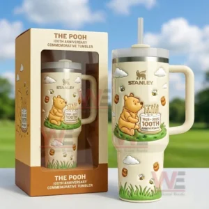 Cream Stanley tumbler with Pooh illustration bees clouds honey pots and 1925 2025 100th anniversary badge shown with a matching commemorative box outdoors