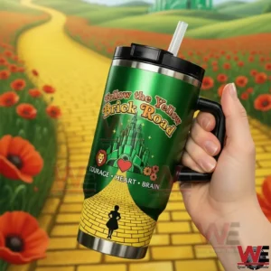 Hand holding a green Follow the Yellow Brick Road tumbler with Emerald City design and courage heart brain icons in front of a yellow brick road and red poppy field!